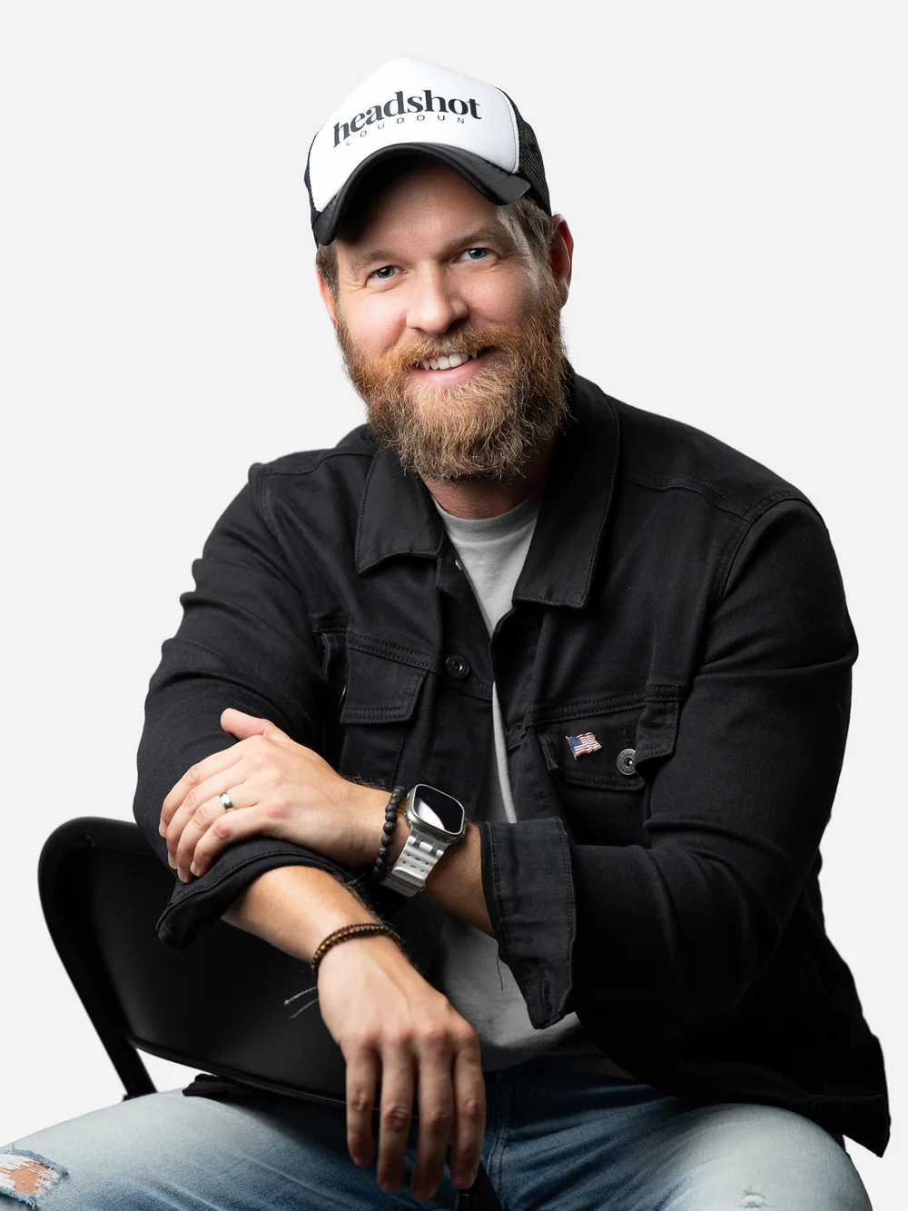 Scott Henderson, smiling, seated on a black chair in a black denim jacket and cap.