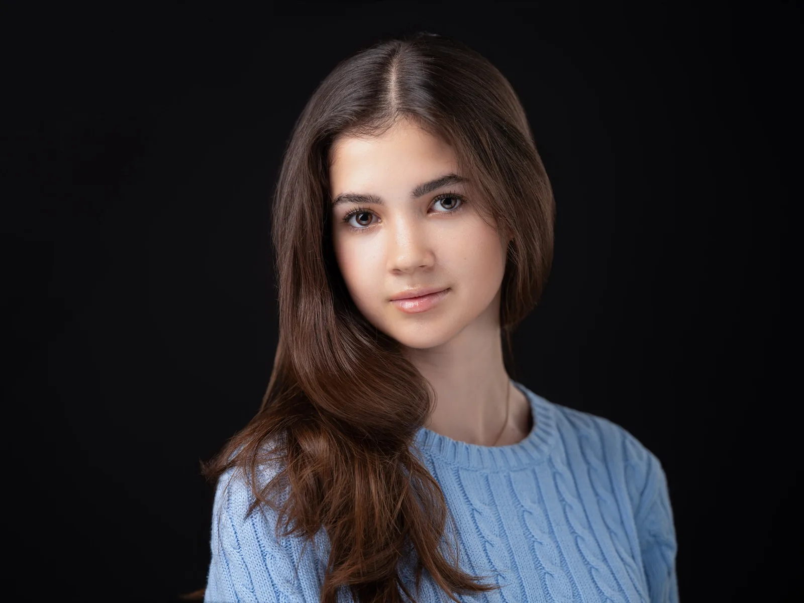 Headshot of a young performer with long brown hair in a light blue sweater, against a black background.